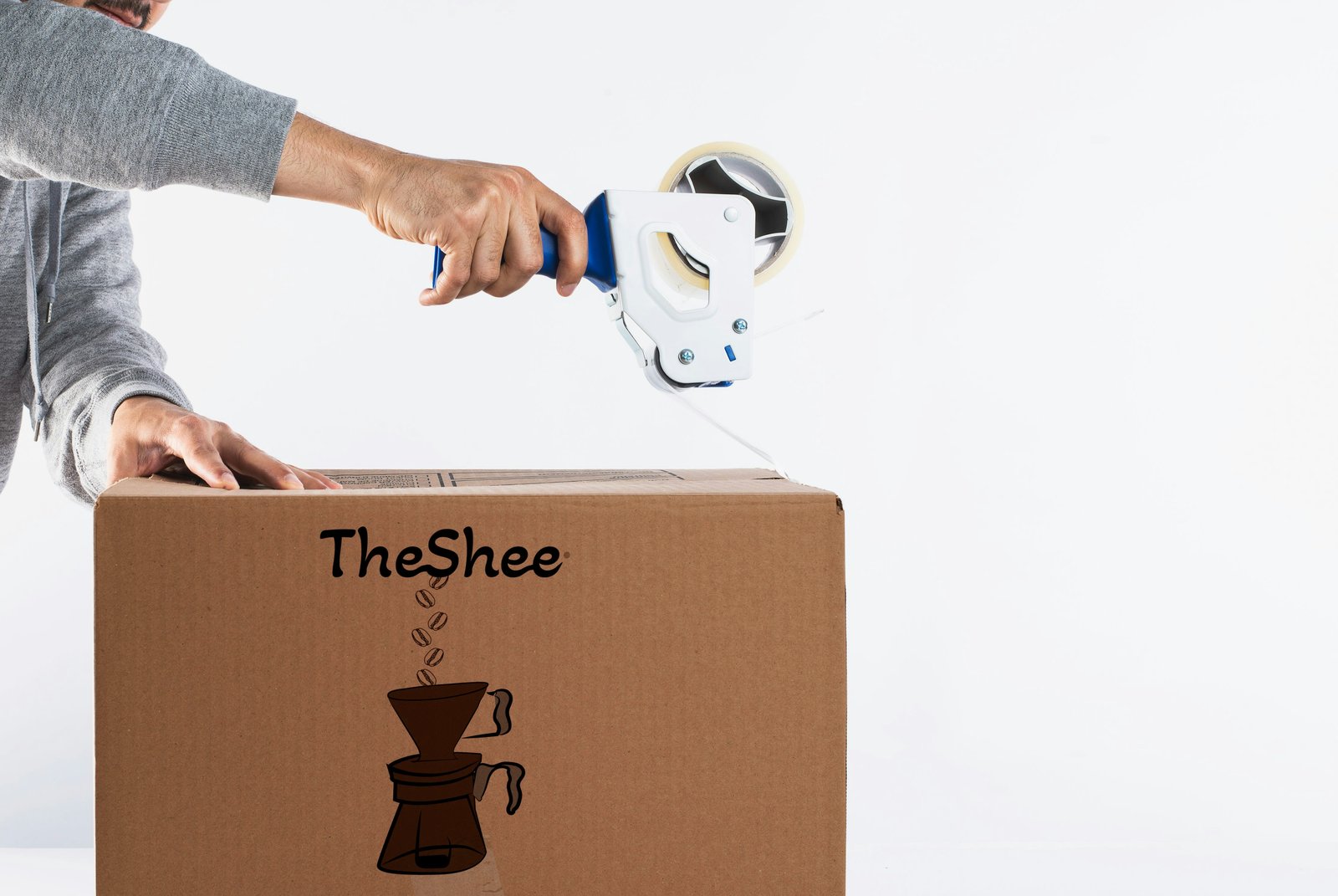 A hand taping a cardboard box printed with the TheShee logo and a line drawing of coffee beans falling into a pour-over dripper